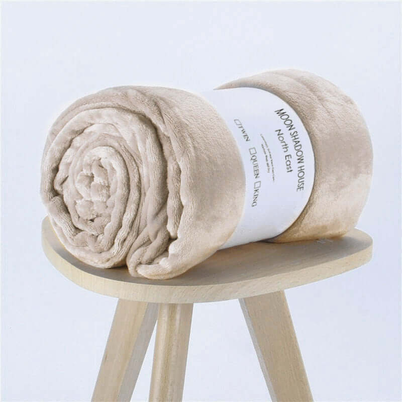Rolled beige flannel coral fleece blanket on a wooden stool with label, perfect for air-conditioning and cozy naps.